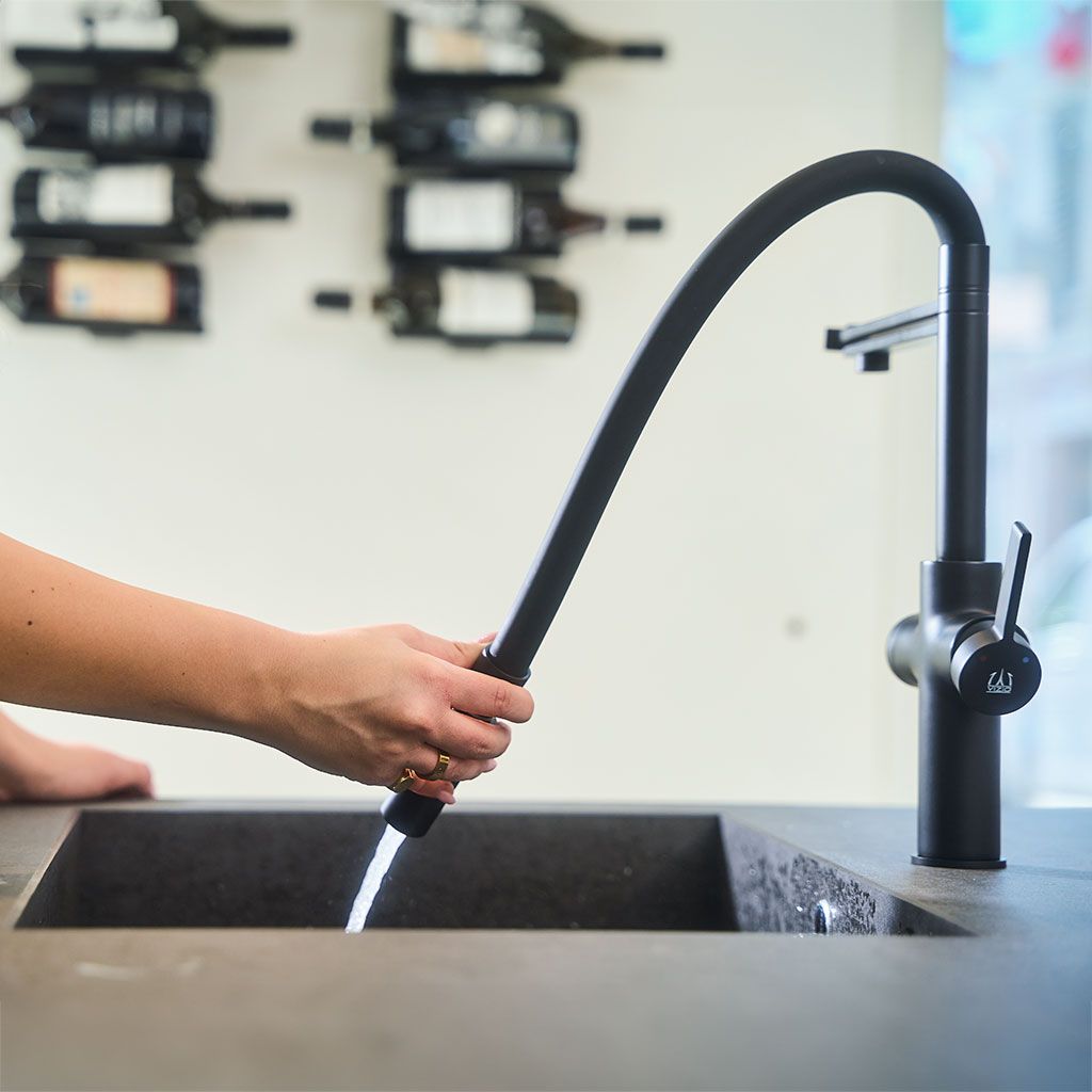 Eine Person hält den Levanto Neo Black 3-Wege-Wasserhahn, während das Wasser in ein schwarzes Waschbecken fließt. Im Hintergrund sind Weinflaschen auf Wandregalen ausgestellt.