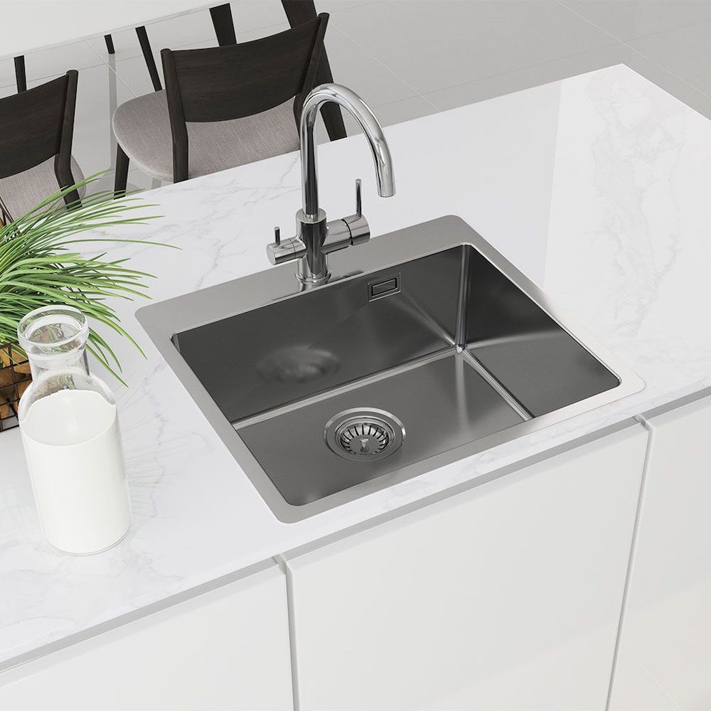 Eine moderne Edelstahlspüle mit dem Basico 3-Wege-Wasserhahn Chrom II ist in eine weiße Marmorarbeitsplatte eingelassen. In der Nähe stehen eine Grünpflanze, ein gläsernes Milchkännchen und eine Holzschüssel, im Hintergrund stehen braune Stühle und ein Esstisch.