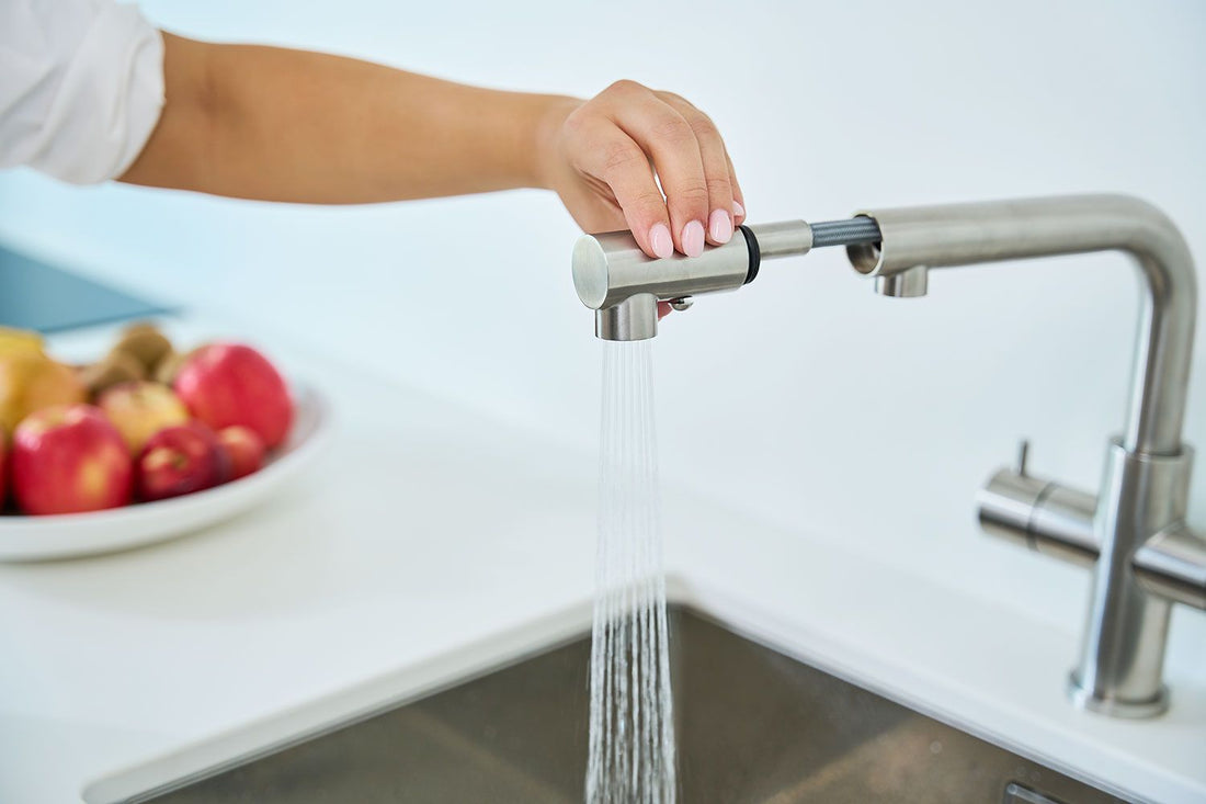 Eine Hand dreht an einem Calabro 3/4-Wege-Wasserhahn Edelstahl mit Ausziehbrause, der Wasser in ein Edelstahlbecken leitet. Im Hintergrund ruht ein weißer Teller mit Äpfeln und Bananen auf einer weißen Arbeitsplatte.