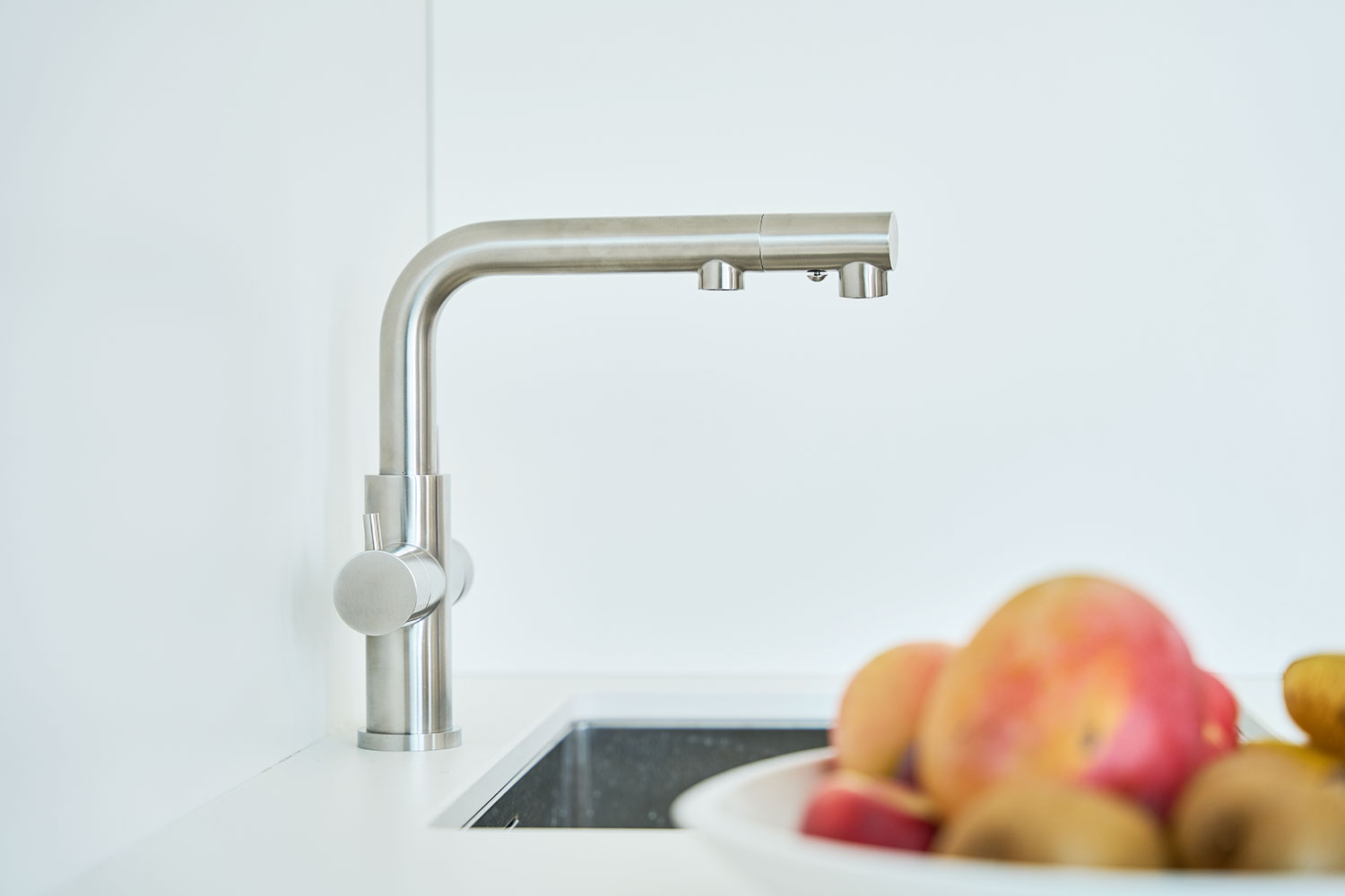 Der Calabro 3/4-Wege-Wasserhahn aus Edelstahl mit Ausziehbrause ist über einem Spülbecken installiert; im Vordergrund steht unscharf eine Obstschale mit Äpfeln vor weißem Hintergrund.