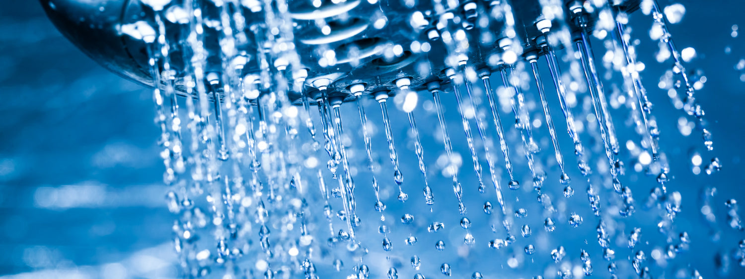 Nahaufnahme von Wassertropfen, die aus einem Duschkopf strömen, wobei das Wasser in der Luft vor einem blau getönten Hintergrund schwebt.
