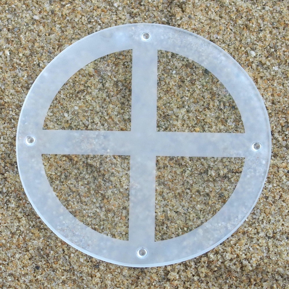 Der UMH Energy 18, eine runde, halbtransparente Siliciumoxid-Scheibe mit einer kreuzförmigen Aussparung und vier Löchern in Randnähe für den Elektrosmog-Schutz, ruht auf einem grob sandigen Untergrund.