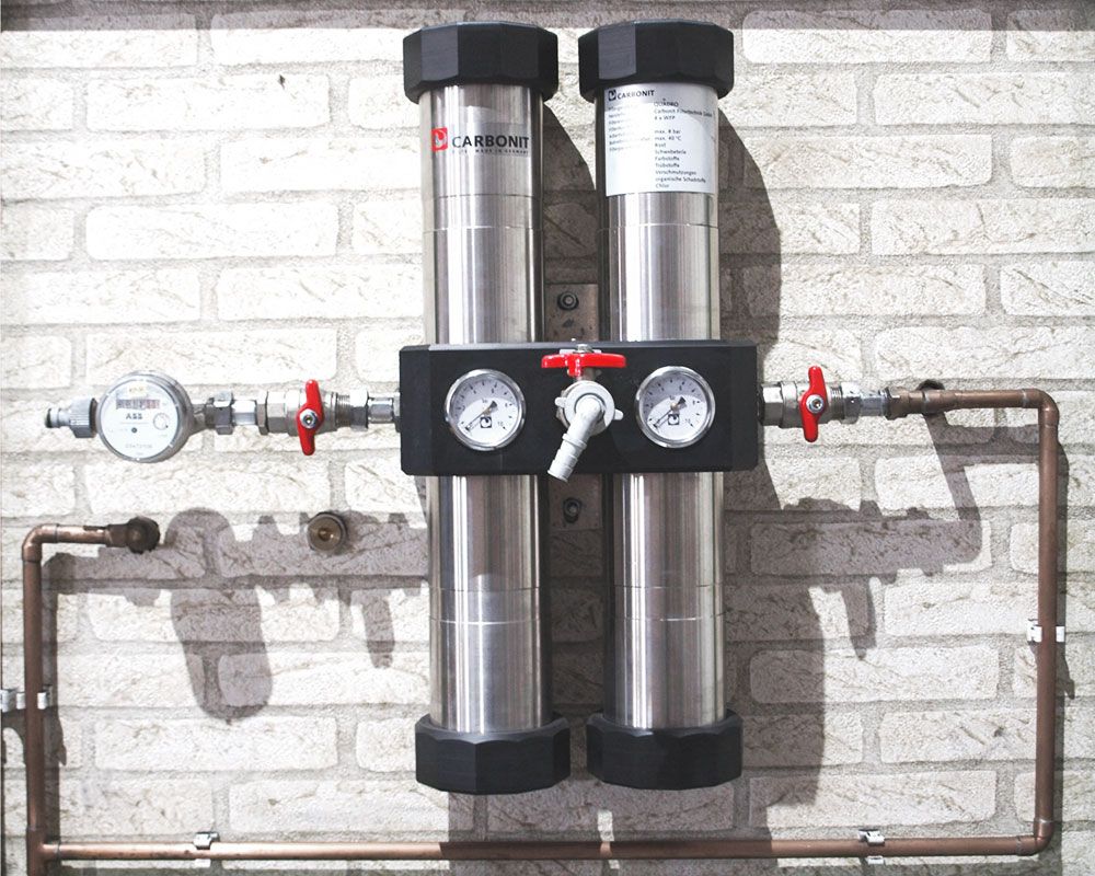 Zwei Carbonit QUADRO Hauswasserfilter-Kanister von Carbonit, jeweils mit Manometer und Rotgriffventil, sind an einer Ziegelwand montiert und mit Kupferrohren verbunden, so dass eine effiziente zentrale Filteranlage zur Wasseraufbereitung entsteht.