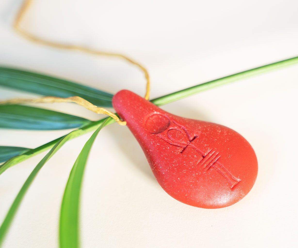 Der Anhänger Stein der Harmonie von Stein der Harmonie besteht aus einem roten Tropfen mit eingraviertem Symbol, der an einer goldenen Kordel hängt und von grünen Palmenblättern getragen wird, die Energie und Ruhe ausstrahlen.