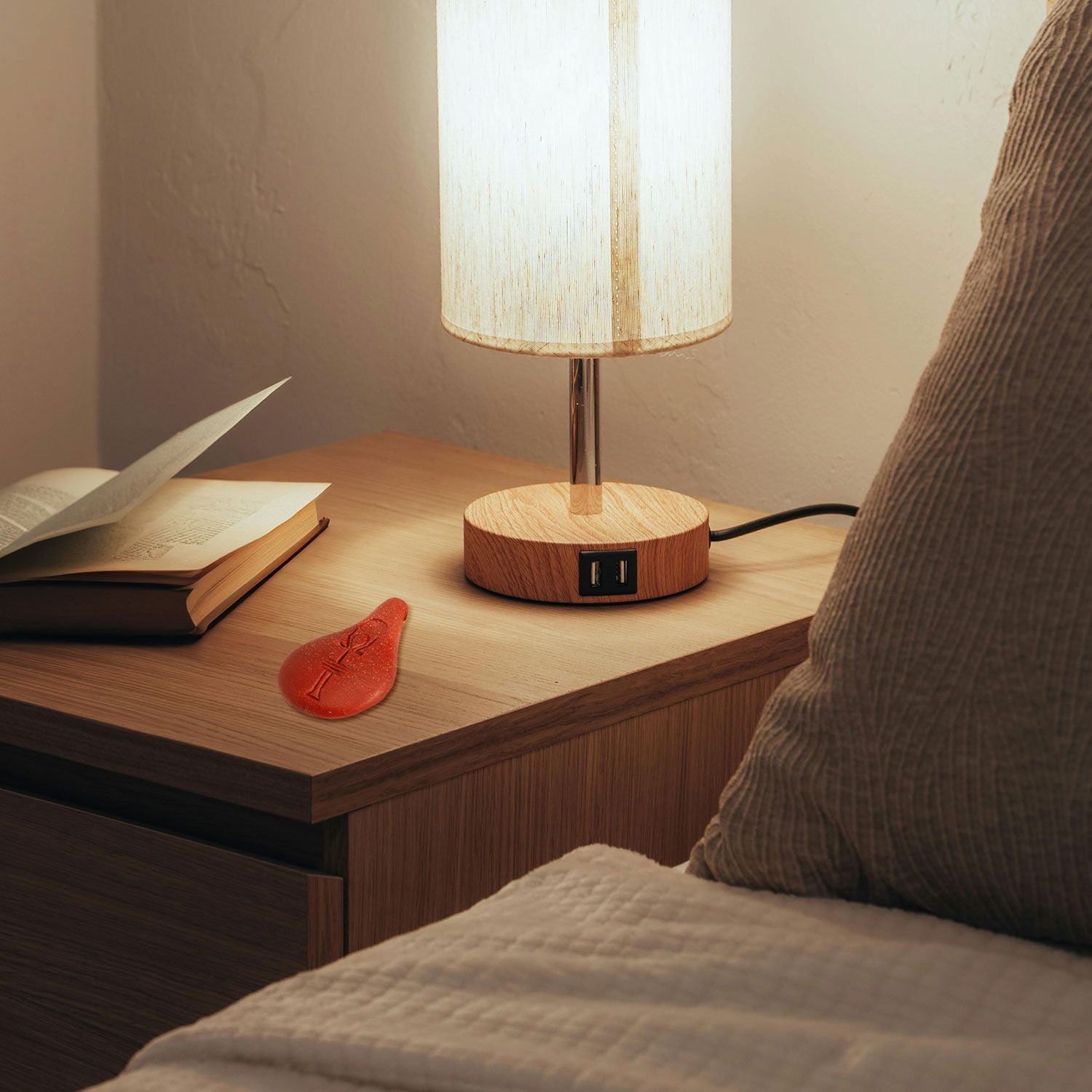 Ein Nachttisch mit einer Holzlampe, ein aufgeschlagenes Buch, ein rotes Lesezeichen und ein Bett mit beigem Bettzeug, das teilweise im Vordergrund zu sehen ist.