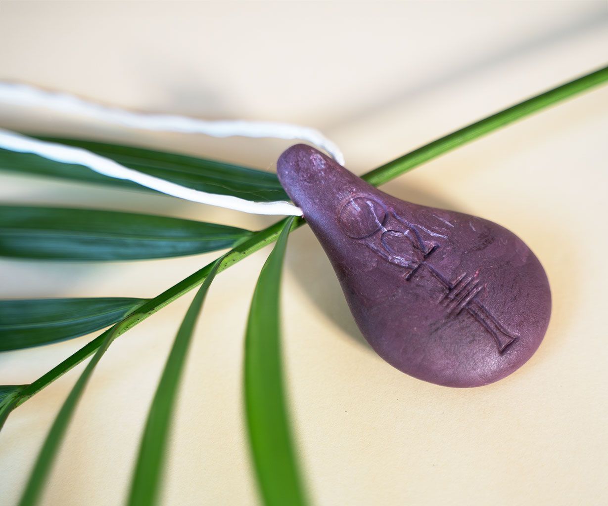 Der Anhänger Stein der Harmonie von Stein der Harmonie zeigt ein violettes Tränenmotiv, in das ein ägyptisches Ankh eingraviert ist. Es ist auf einem grünen Palmenblatt angebracht und hängt an einer weißen, gedrehten Schnur vor einem hellen Hintergrund.