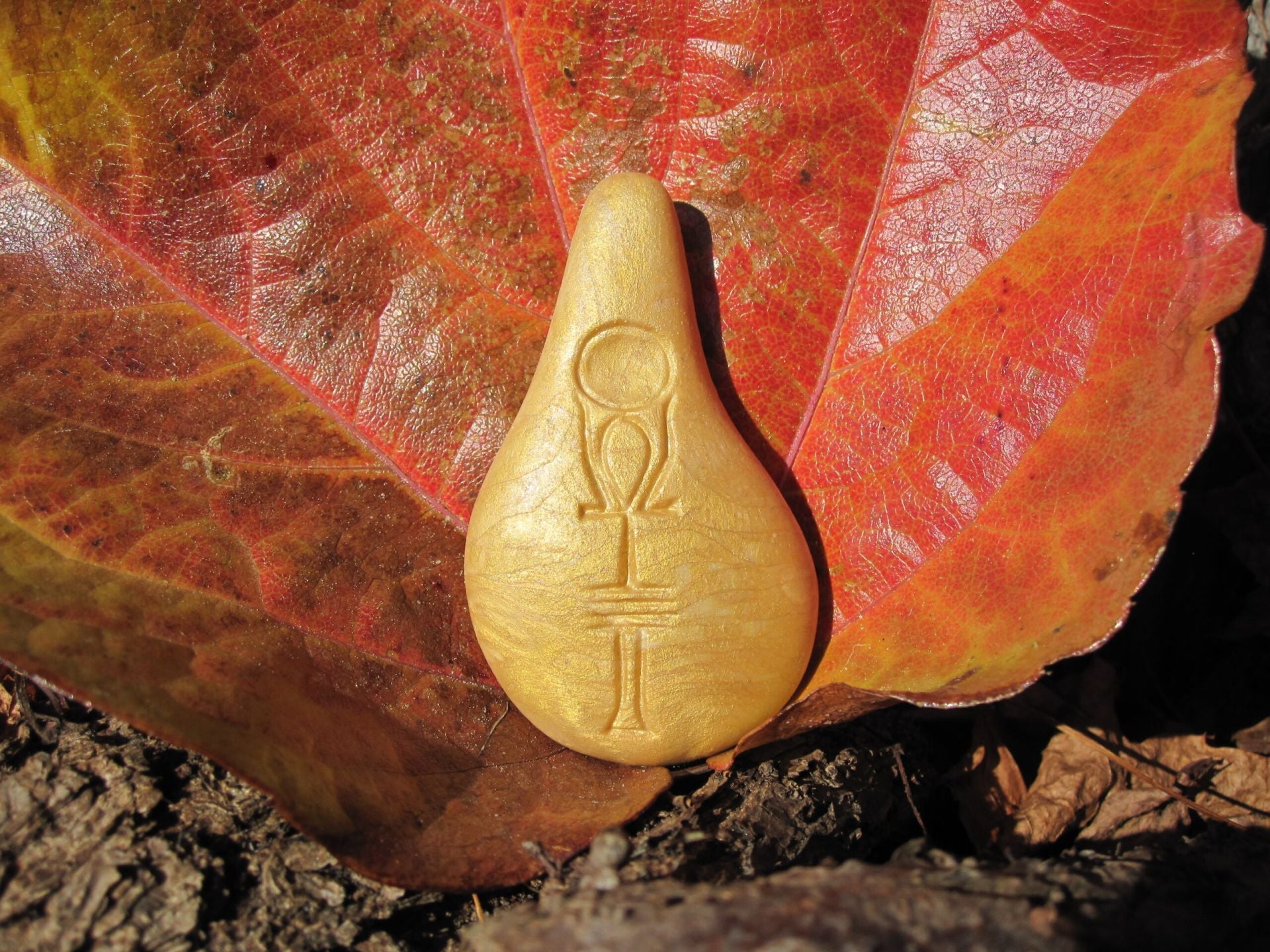 Ein hellbrauner, tropfenförmiger Stein der Harmonie von Stein der Harmonie ruht auf einem großen roten und orangefarbenen Blatt im Freien. Mit Symbolen graviert, strahlt er Energie aus und zeigt ein Omega über einem Kreuz mit zwei horizontalen Linien.