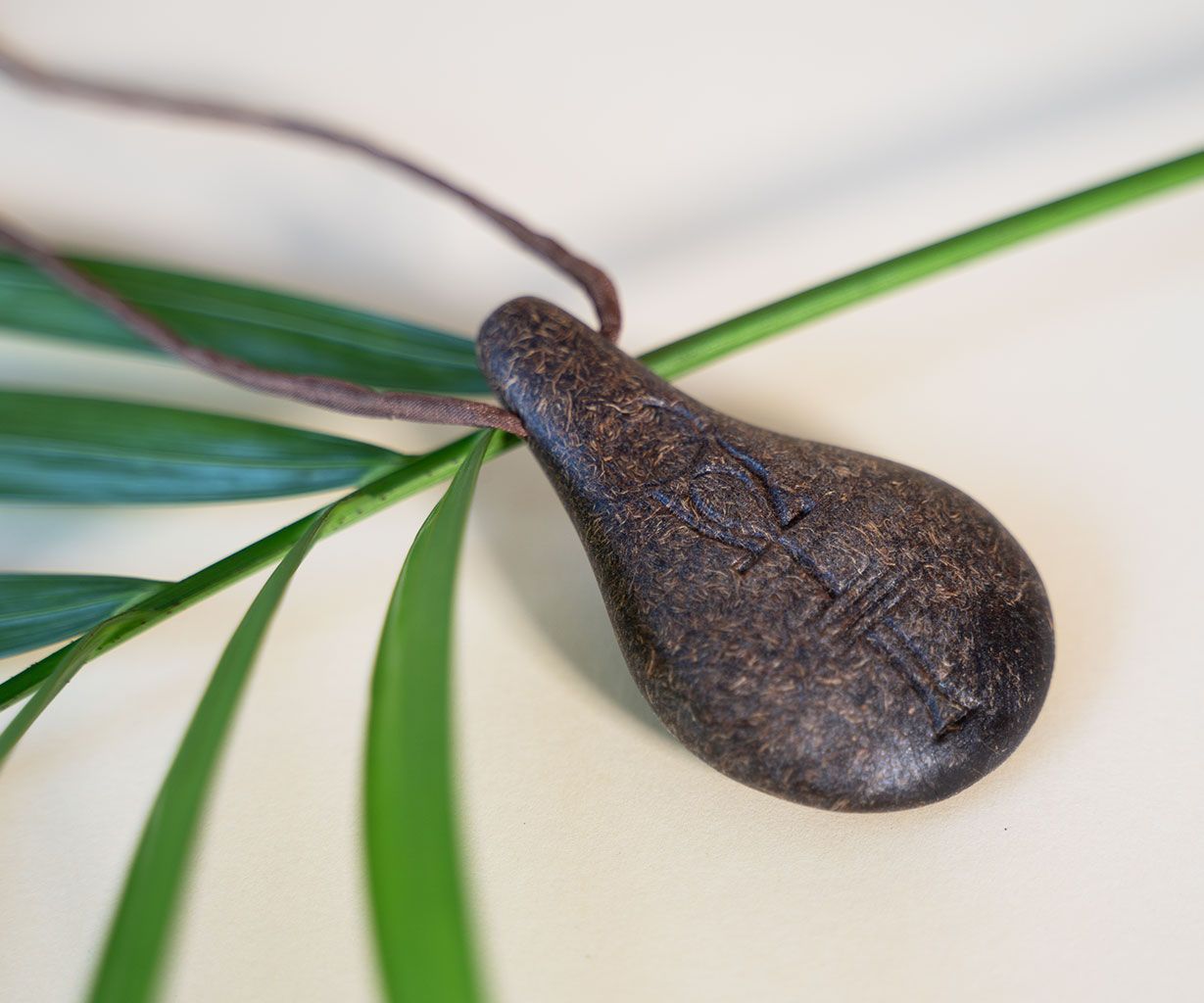 Der Anhänger Stein der Harmonie von Stein der Harmonie, eine dunkle Tropfenform mit Gravuren, hängt an einer braunen Kordel auf einer hellen Fläche mit grünen Blättern - ein Symbol für innere Balance und positive Energie.