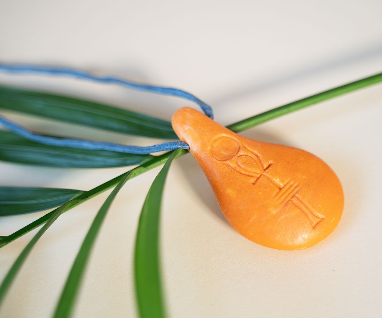 Der Stein der Harmonie von Stein der Harmonie ist ein orangefarbener, tropfenförmiger Anhänger mit symbolischen Gravuren, der an einer blauen Schnur über grünen Palmenblättern hängt. Er soll die innere Balance und positive Energie vor einem hellen Hintergrund fördern.