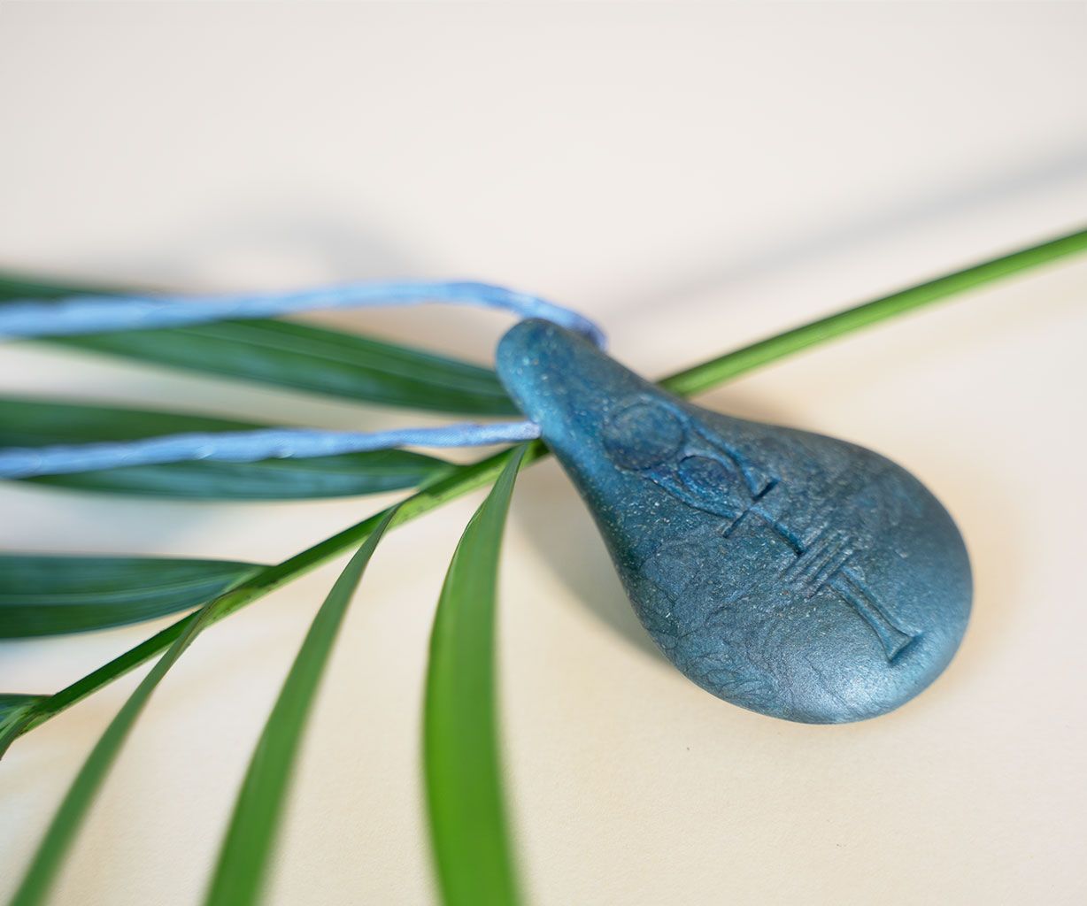 Ein Stein der Harmonie-Anhänger von Stein der Harmonie mit einem blauen Stein mit eingraviertem Ankh-Symbol ruht auf einem Palmblatt mit einer blauen Kordel - er soll Energie ausstrahlen und die innere Balance unterstützen.