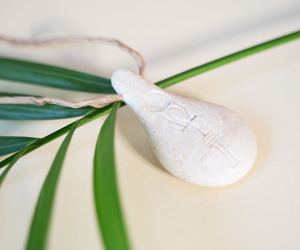 Der Stein der Harmonie von Stein der Harmonie ist ein weißer, tropfenförmiger Anhänger mit geprägtem Design an einer hellbraunen Kordel, die auf grünen Palmenblättern vor einem hellen Hintergrund steht.