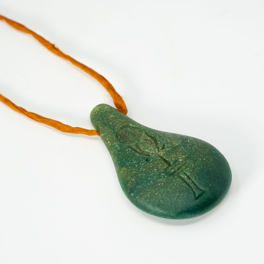 Der Anhänger "Stein der Harmonie" von Stein der Harmonie hat die Form einer grünen Träne und ein eingraviertes Symbol, das das innere Gleichgewicht und die positive Energie fördert. Er hängt an einer braunen Kordel und ist auf einem weißen Hintergrund abgebildet.