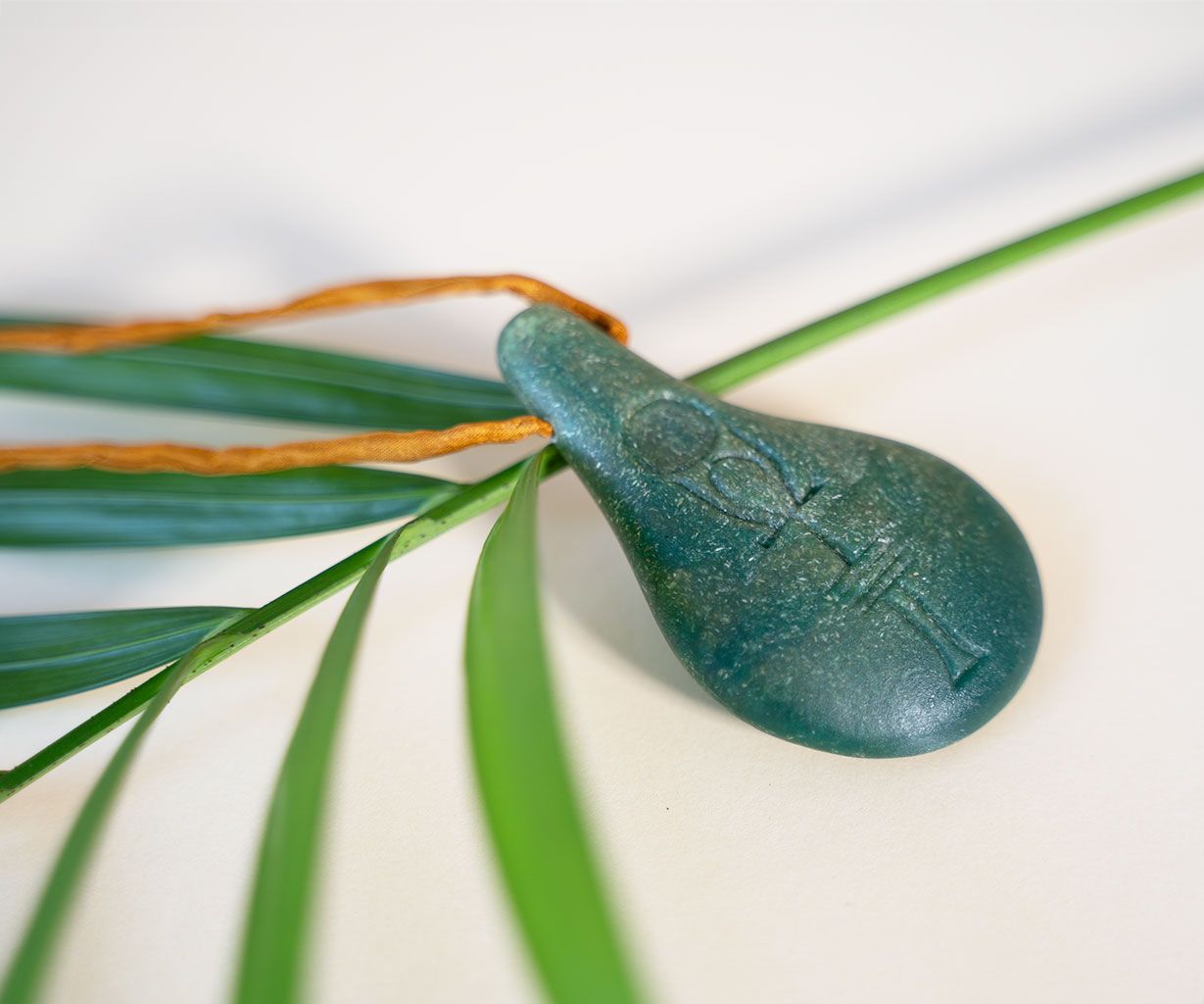 Der Stein der Harmonie von Stein der Harmonie ist ein tropfenförmiger Anhänger aus Grünstein mit eingravierten Markierungen und einer Schnur, die auf Palmenblättern ruht. Er symbolisiert das innere Gleichgewicht vor einem sanft verschwommenen Hintergrund.