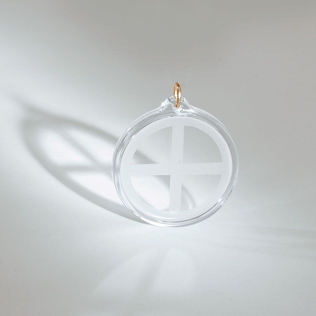 Der UMH Energy 45 ist ein klarer, runder Elektrosmog-Schutz-Anhänger mit weißem Kreuzmotiv, der aufrecht auf einer weißen Fläche steht und einen Schatten wirft. Er verfügt über eine kleine goldene Schlaufe an der Oberseite, um ihn an einer Kette zu befestigen.