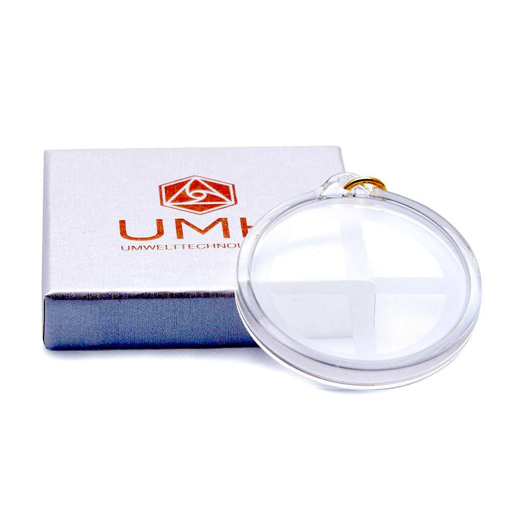 Ein runder, klarer UMH Energy 45-Anhänger mit Kreuzmotiv, der an einem goldenen Ring befestigt ist, befindet sich neben einer grauen Box mit der Aufschrift UMH UMWELTTECHNOLOGIE und einem roten Elektrosmog Schutz-Logo.