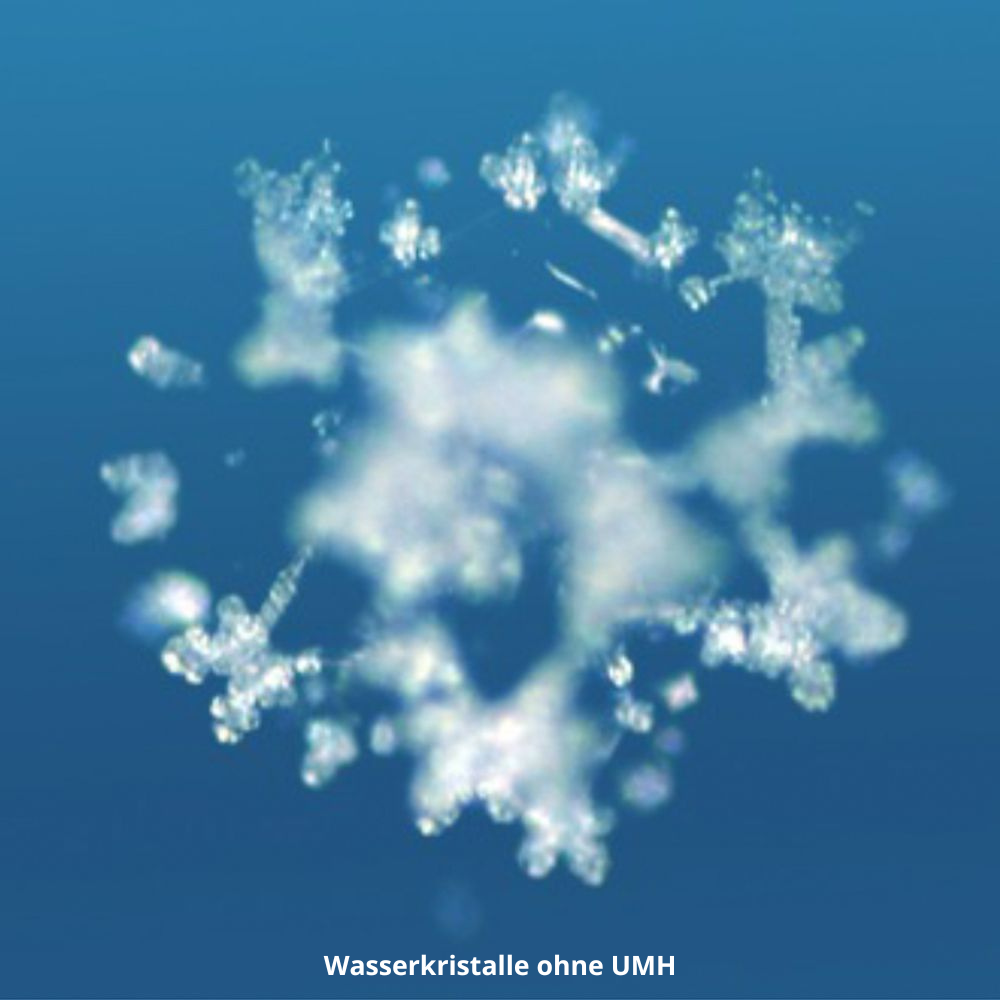 Ein verschwommener, weißer, schneeflockenartiger Kristall bildet sich vor einem blauen Hintergrund. Der Text darunter lautet "Wasserkristalle ohne UMH" und zeigt einen Wasserkristall ohne UMH PURE GOLD Wasser-Energetisierung von UMH.
