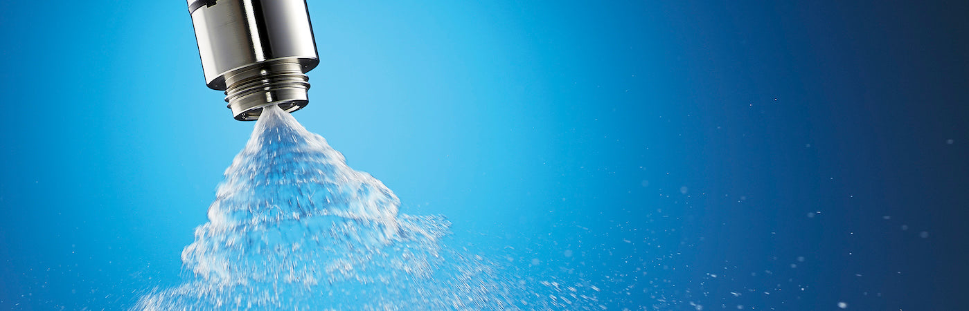 Nahaufnahme eines Sprinklerkopfes aus Metall, der Wasser in einem feinen Nebel vor einem blauen Hintergrund mit Farbverlauf versprüht.