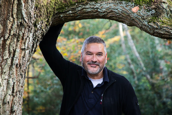 Ein Mann mit kurzen grauen Haaren und einem Bart lächelt in die Kamera, während er im Freien steht und seinen Arm auf einen gebogenen Baumzweig stützt. Im Hintergrund ist üppiges grünes Laub zu sehen.