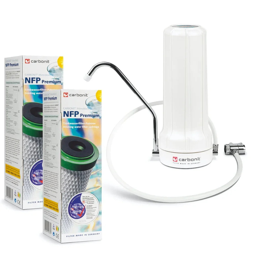 Ein Carbonit SANUNO Auftischfilter mit Metalltülle, Schlauch und Anschlussstück steht neben zwei Kartons mit NFP Premium Ersatzkartuschen. Auf der Verpackung sind Bilder des Filters und von Wassertropfen abgebildet.
