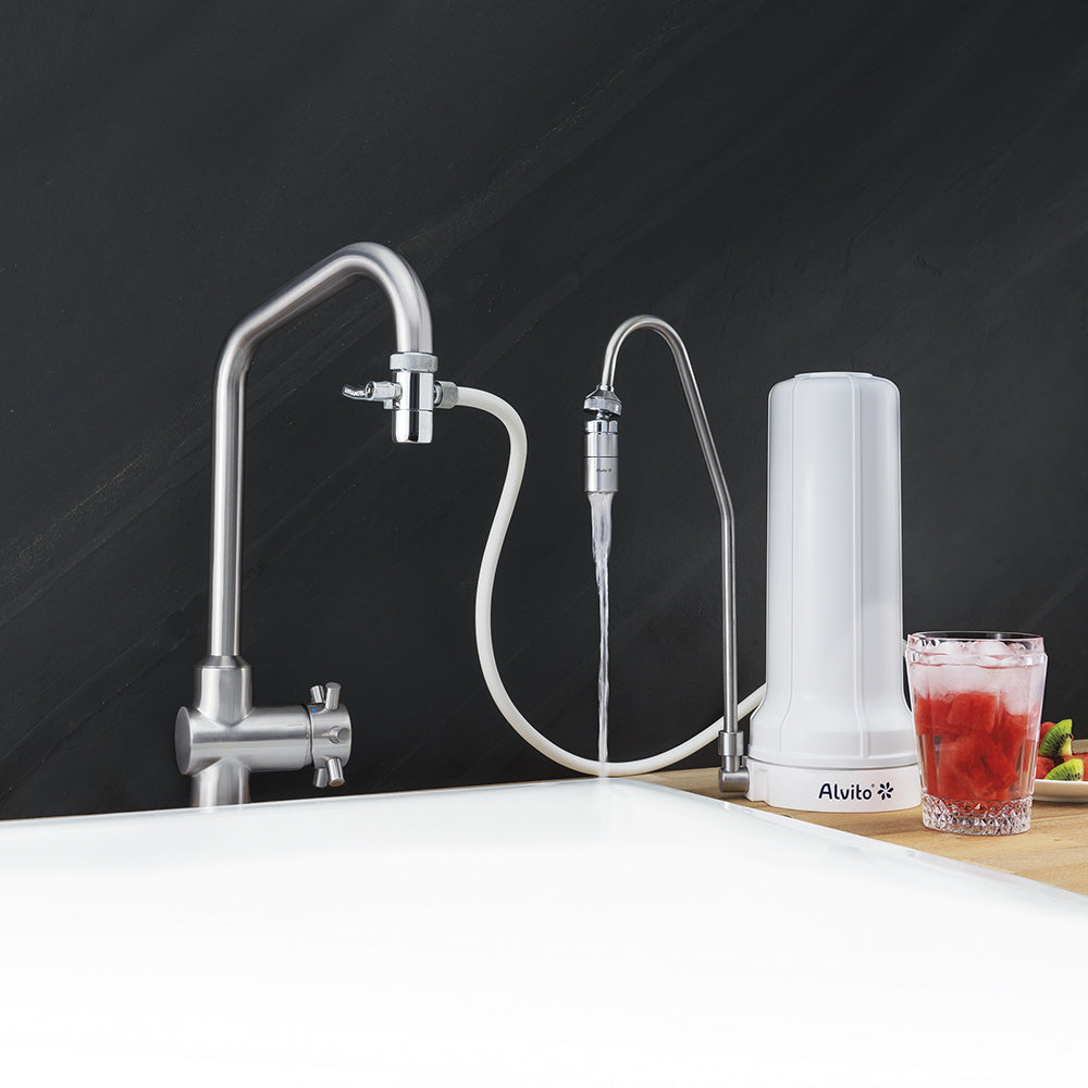 Das Alvito Auftischfilter Set Natur mit Aktivkohle-Filter spendet Wasser in ein Waschbecken. Daneben steht ein Glas rotes Getränk mit Eis auf einer Holztheke.