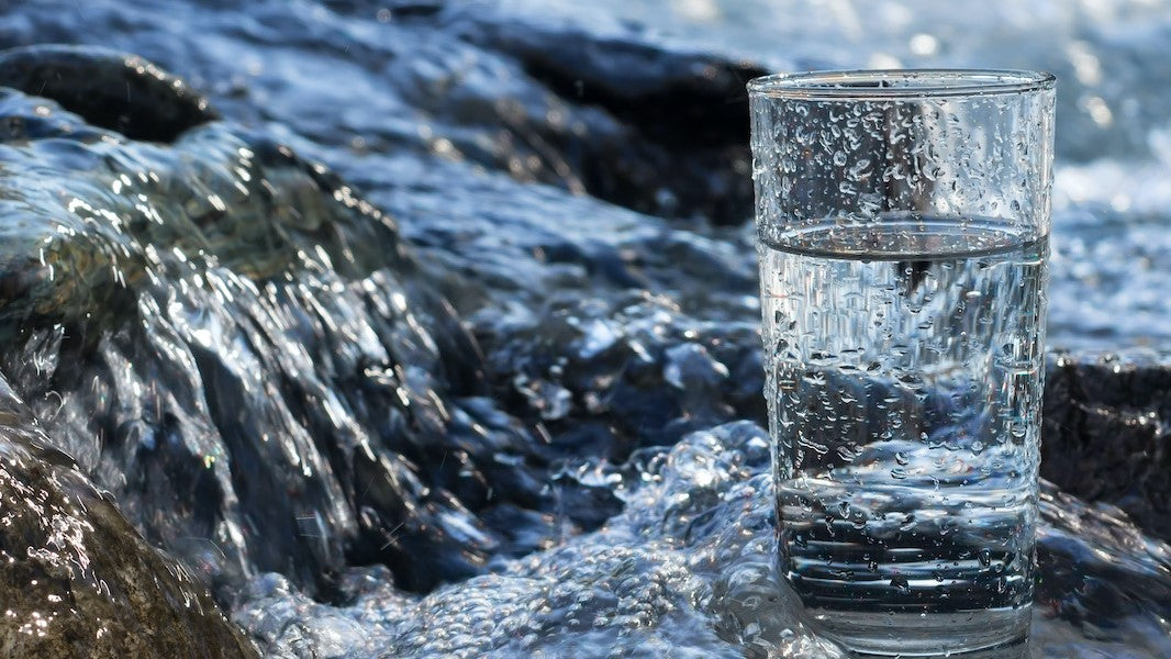 Glas mit hexagonalem Wasser auf Felsen mit fließendem Bach im Hintergrund