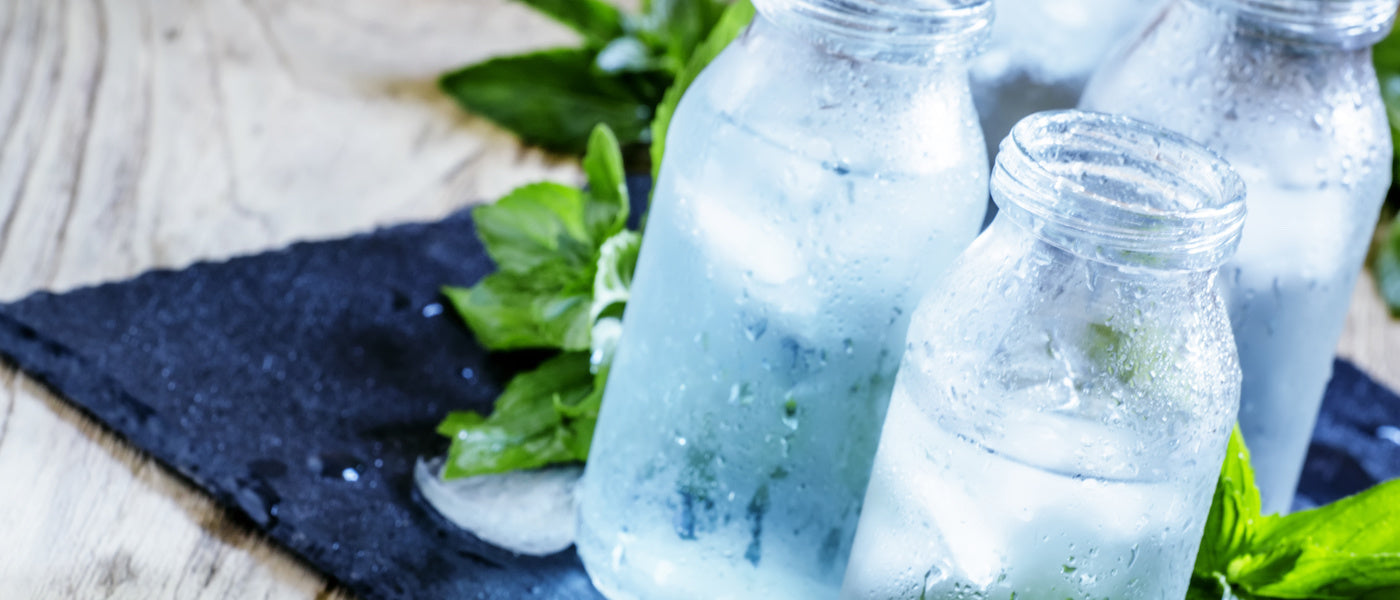 Drei mit Eiswasser gefüllte Eisgläser stehen auf einer dunklen Schieferfläche, umgeben von frischen grünen Minzblättern, an denen sich Kondenswasser bildet. Der Hintergrund ist ein heller Holztisch.