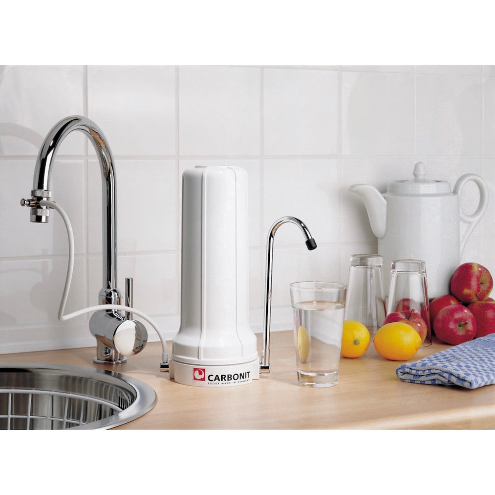 Auf einer Küchenarbeitsplatte stehen ein Carbonit SANUNO Wasserfilter von Carbonit, ein Waschbecken mit einem am Wasserhahn montierten Filter, ein Glas Wasser, Obst, zwei umgedrehte Gläser, eine Teekanne und ein gefaltetes blaues Handtuch vor weiß gekachelten Wänden.