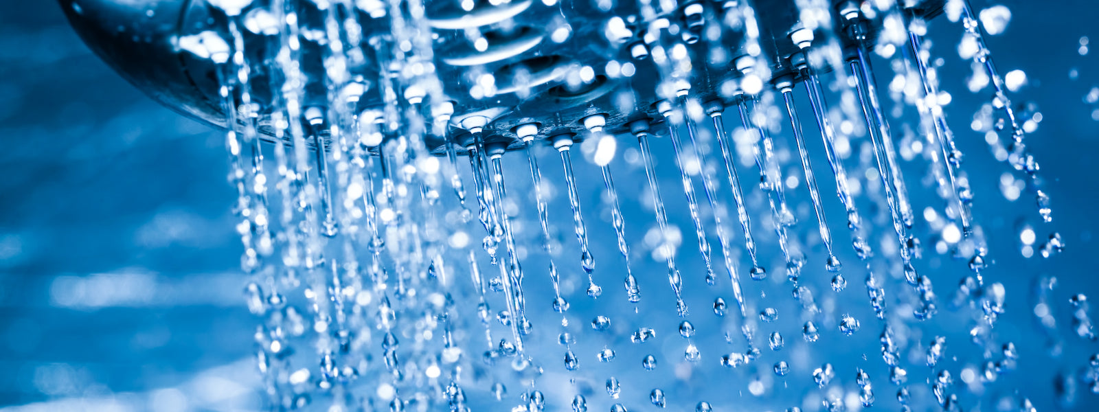 Nahaufnahme von Wassertropfen, die aus einem Duschkopf strömen, wobei das Wasser in der Luft vor einem blau getönten Hintergrund schwebt.