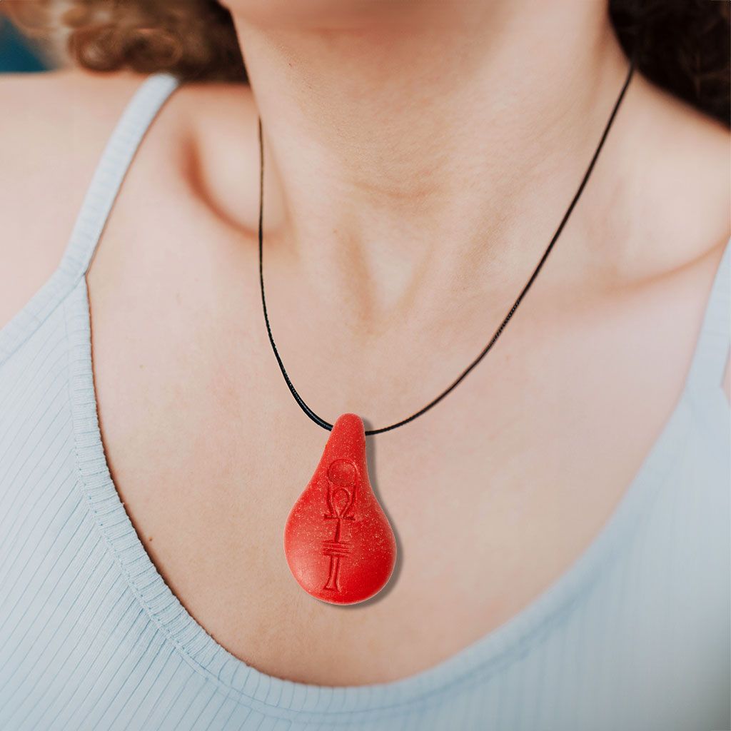 Eine Person, die ein hellblaues, geripptes Tank-Top und eine Halskette mit einem großen roten, tropfenförmigen Anhänger mit einem eingravierten Ankh-Symbol trägt.